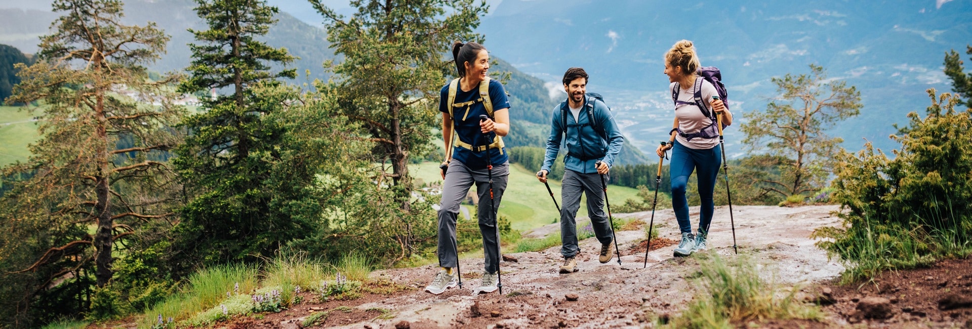 Drei Wanderer mit Rucksäcken und Wanderstöcken genießen einen Bergpfad mit Blick auf grüne Hügel und Bäume. Sie lächeln und scheinen eine gute Zeit zu haben.