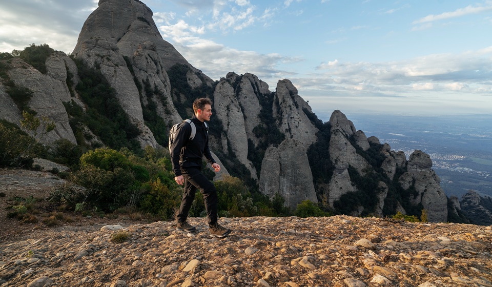 A man is hiking on a mountain peak, behind him stretch rock formations and a vast landscape.