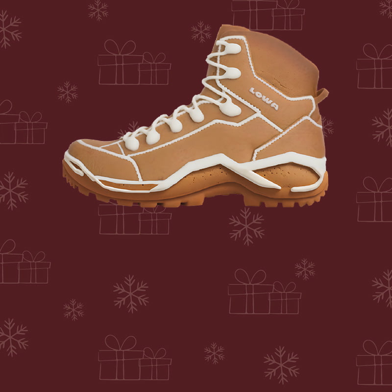 The picture shows a gingerbread shoe from the brand Lowa in front of a dark red background with a snowflake and gift pattern.