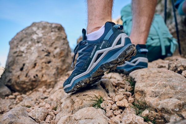 Wanderer mit blauen Laufschuhen auf felsigem Gelände. Die Schuhe bieten guten Halt. Im Hintergrund ein Rucksack. Sonniger Tag.