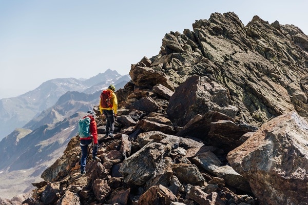 Zwei Wanderer mit Rucksäcken erklimmen einen felsigen Berggrat. Die atemberaubende Berglandschaft erstreckt sich weitläufig im Hintergrund. Ein sonniger Tag mit klarem Himmel.