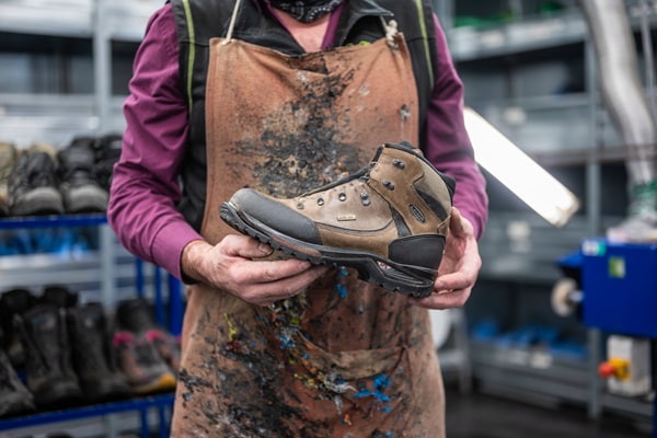 Ein Schuhmacher präsentiert einen robusten, braunen Wanderschuh. Seine schmutzbefleckte Schürze und der Hintergrund mit Schuhregalen zeigen seine Werkstatt. Der Fokus liegt auf dem Schuh in seinen Händen.