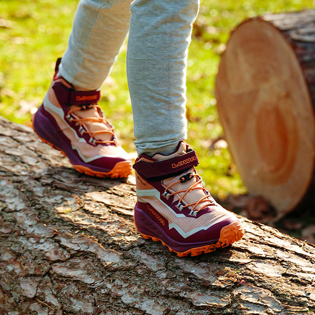 Kind in leuchtend orangen Wanderschuhe steht auf einem Baumstamm in einer natürlichen Umgebung.