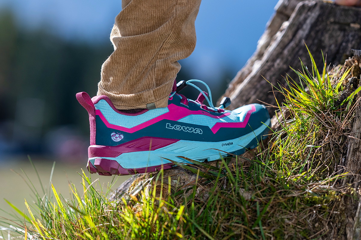 Fotografie zeigt ein Kind, das blau-pinke Wanderschuhe mit weißen Details auf einem steinigen Boden trägt. Fokus liegt auf Schuhen, während unscharfer Hintergrund Naturumgebung andeutet.