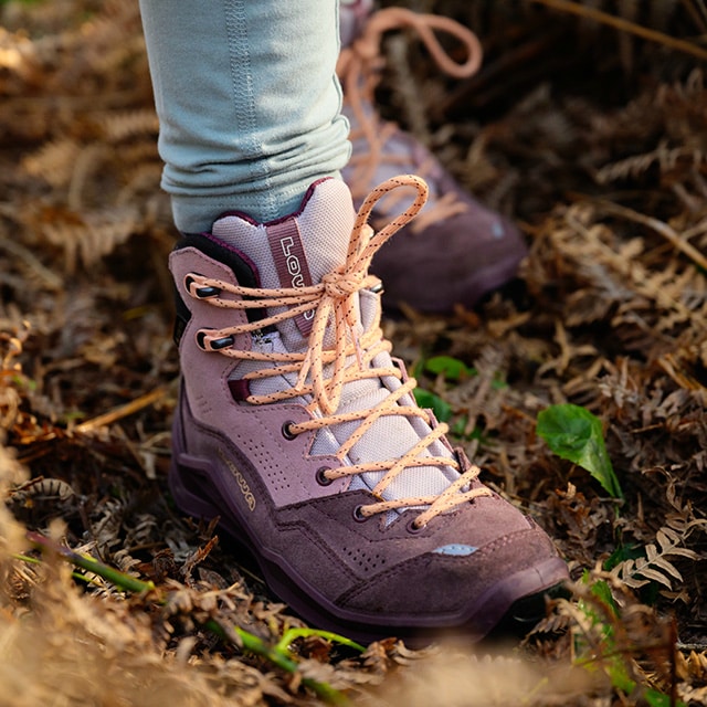 A child in bright orange hiking boots stands on a tree trunk in a natural setting.