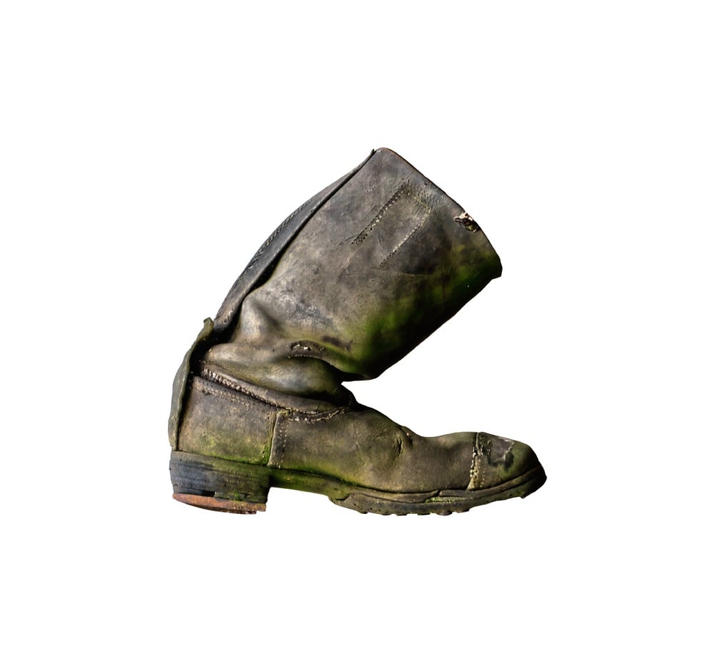 Ein alter, abgenutzter, grün bewachsener Lederstiefel liegt auf dem Kopf. Der Stiefel ist stark verschmutzt und weist Beschädigungen auf. Der Hintergrund ist weiß.