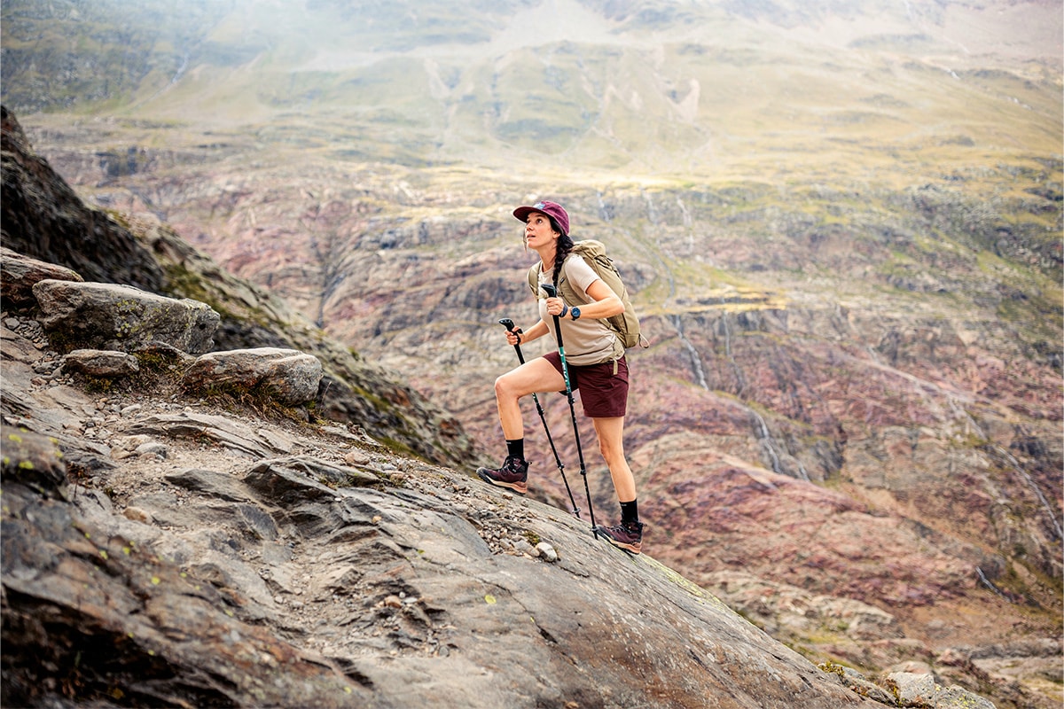 Fotografie einer Wanderin, die mit Trekkingstöcken einen steilen, felsigen Bergpfad erklimmt, umgeben von einer weitläufigen, gebirgigen Landschaft. Die Person trägt Outdoor-Bekleidung, Rucksack und Wanderschuhe, was auf eine anspruchsvolle Bergtour hinweist.