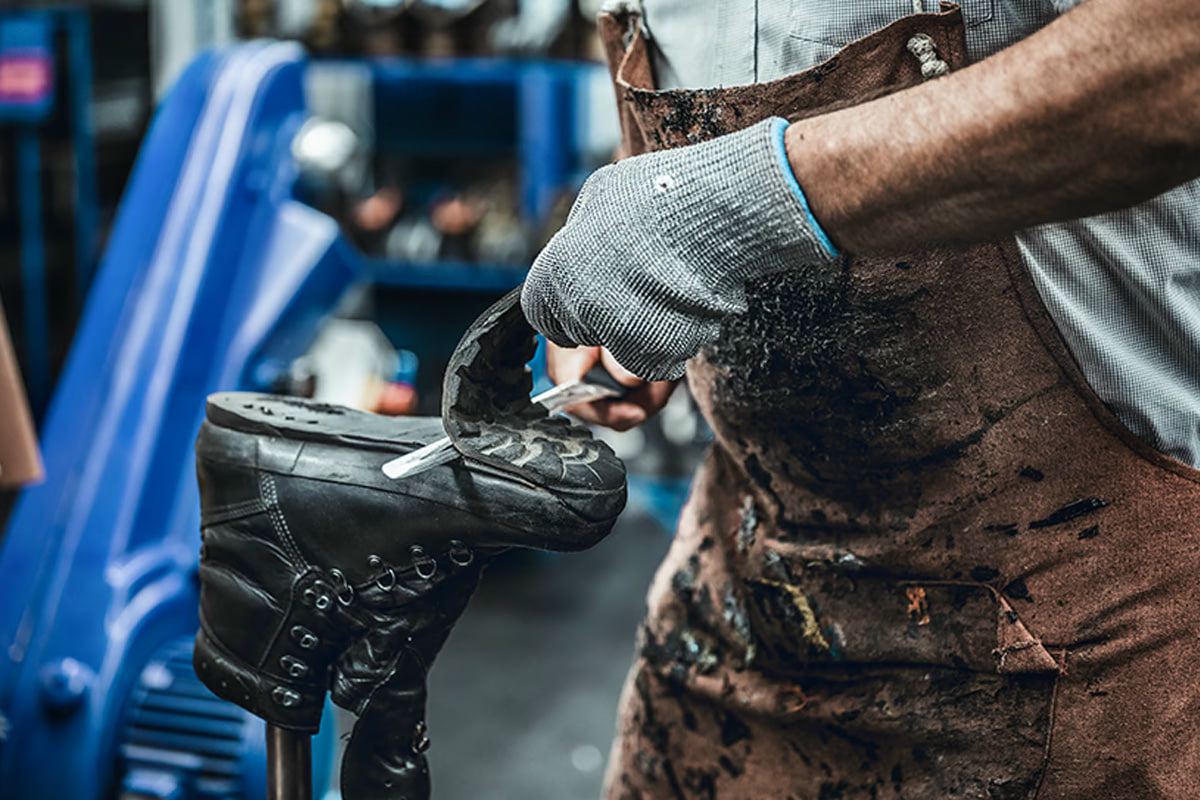Fotografie eines Schuhmachers bei der Arbeit, der mit Handschuhen einen schwarzen Stiefel repariert. Fokus auf Handwerk und Details wie Werkzeug, abgenutzte Lederfläche und schmutzige Schürze.