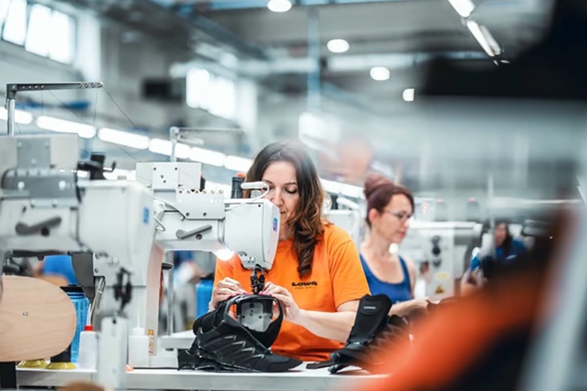 Fotografie einer Produktionslinie in einer Fabrik, auf der mehrere Personen an Nähmaschinen arbeiten, um Schuhe herzustellen. Fokus liegt auf einer Frau in orangefarbenem Shirt, die ein Schuhoberteil bearbeitet, umgeben von industriellen Maschinen und Arbeitsplätzen.