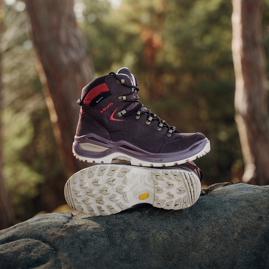 Ein Paar bordeauxrote Wanderschuhe liegt auf einem mit Wurzeln übersäten Waldboden; im Hintergrund schlängelt sich ein verschwommener Pfad durch hohe Bäume.