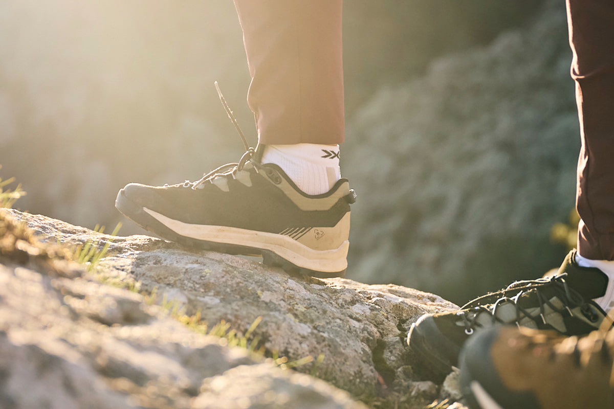 Fotografie zeigt eine Person, die auf einem Felsen sitzt und schwarze Wanderschuhe schnürt, umgeben von Berglandschaft mit unscharfem Hintergrund. Fokus liegt auf robusten Schuhen mit stabiler Sohle und Schnürsystem, geeignet für Outdoor-Aktivitäten und Wanderungen.