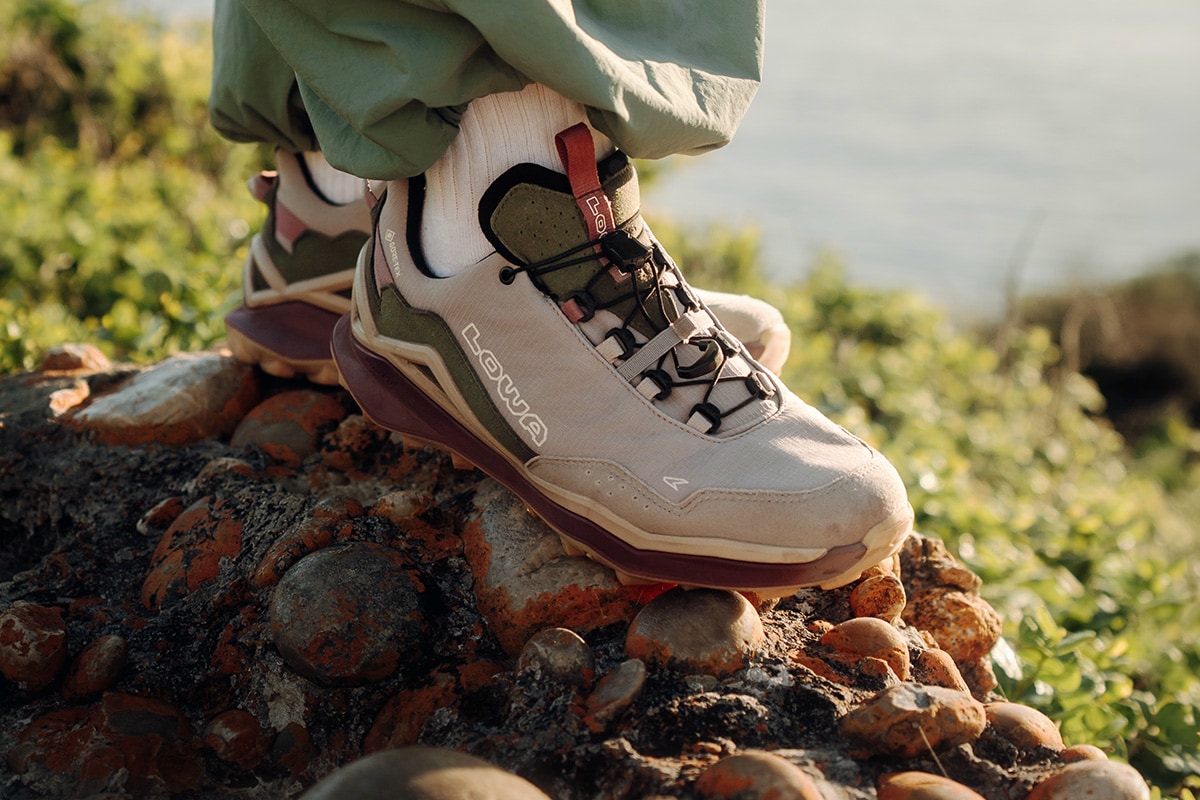 Nahaufnahme eines hellgrauen MADDOX PRO GTX LO SL der Marke Lowa auf felsigem Untergrund mit grüner Vegetation im Hintergrund. Fokus liegt auf robustem Design, stabiler Sohle und Schnürsystem, geeignet für Outdoor-Aktivitäten.