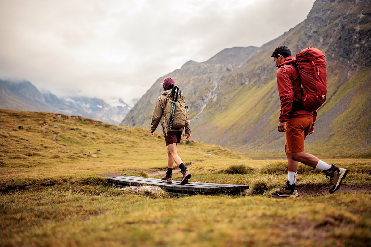 Fotografie von zwei Wanderern mit Rucksäcken, die auf einem Pfad durch eine bergige Landschaft mit bewölktem Himmel gehen. Fokus liegt auf Outdoor-Aktivität und Naturerlebnis, mit grünen Wiesen und steilen Berghängen im Hintergrund.
