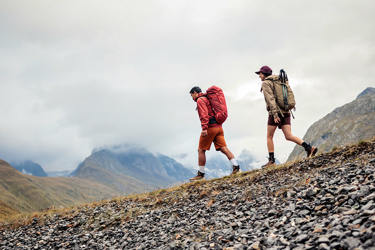 Fotografie zeigt drei Wanderer auf einer grünen Wiese vor bewaldeten Hügeln und felsigen Bergen im Hintergrund. Szene vermittelt Outdoor-Aktivität und Naturerlebnis bei sonnigem Wetter.