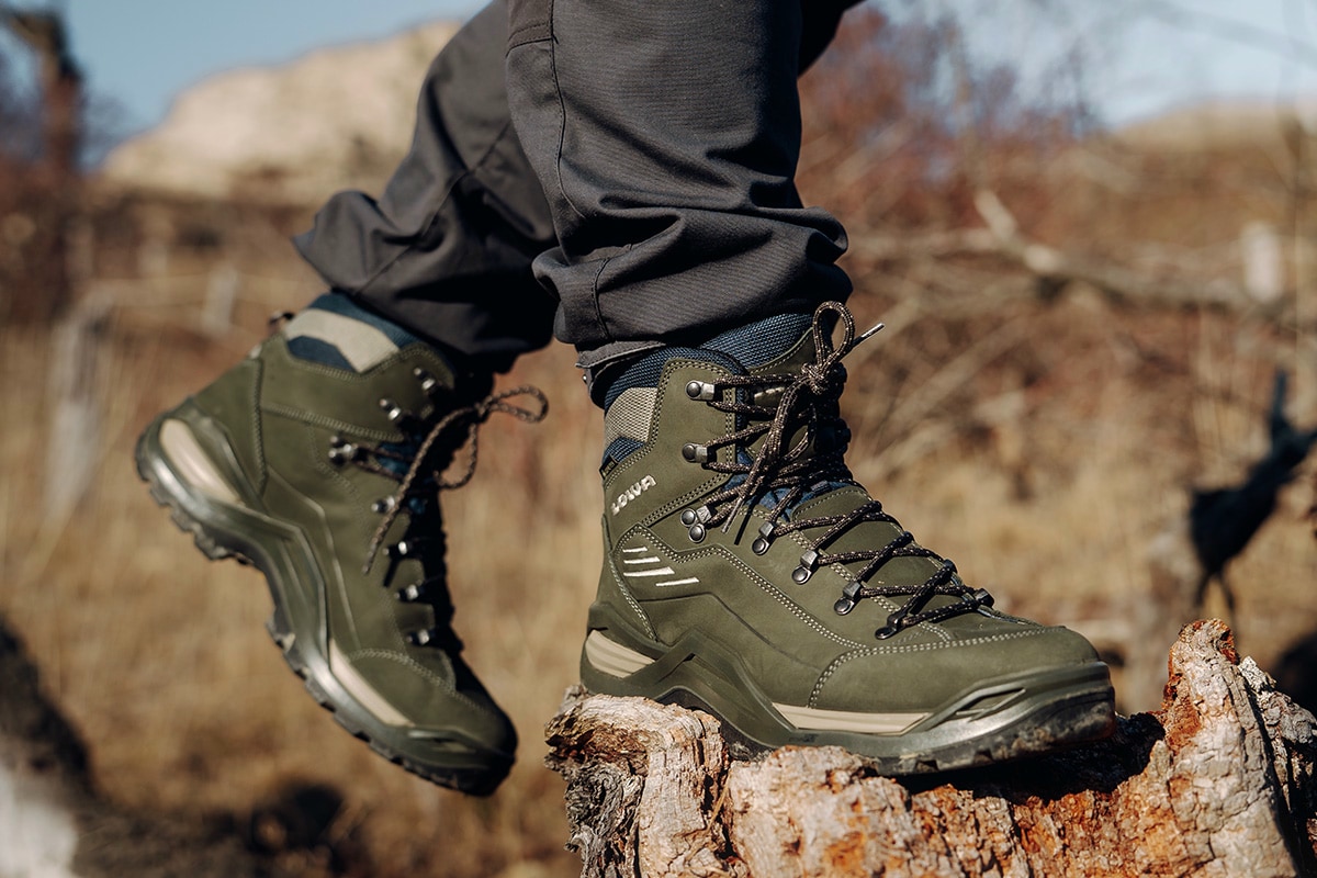 Fotografie zeigt eine Person, die auf einem Felsen sitzt und schwarze Wanderschuhe schnürt, umgeben von Berglandschaft mit unscharfem Hintergrund. Fokus liegt auf robusten Schuhen mit stabiler Sohle und Schnürsystem, geeignet für Outdoor-Aktivitäten und Wanderungen.