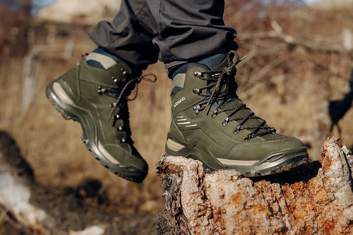 Fotografie zeigt eine Person, die auf einem Felsen sitzt und schwarze Wanderschuhe schnürt, umgeben von Berglandschaft mit unscharfem Hintergrund. Fokus liegt auf robusten Schuhen mit stabiler Sohle und Schnürsystem, geeignet für Outdoor-Aktivitäten und Wanderungen.