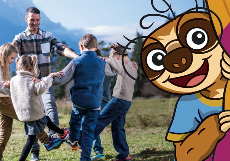 Een groep kinderen en een volwassene staan in een kring op het grasveld en houden elkaars handen vast, terwijl een tekenfilmfiguur in de vorm van een luiaard van rechts naar hen kijkt.