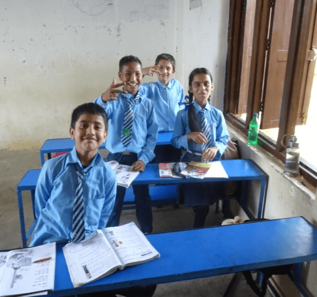 Four students in blue shirts and ties stand smiling behind their tables.