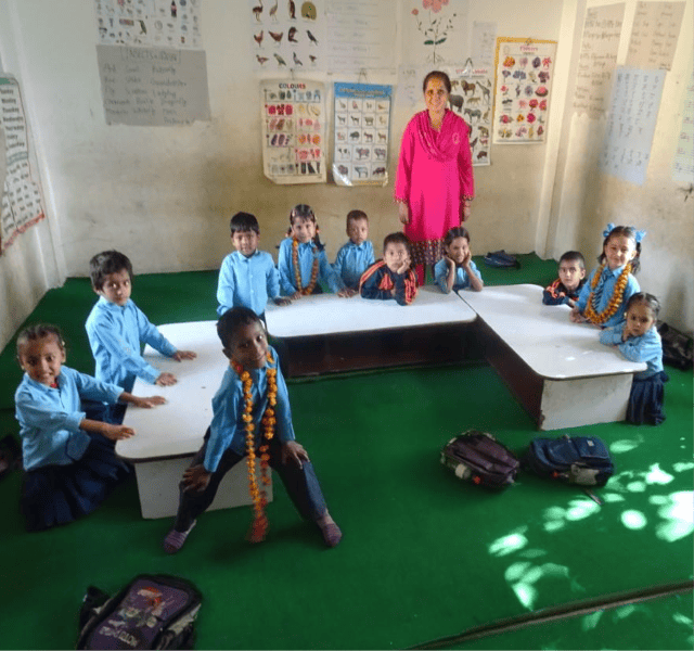 Teacher in pink stands in front of a group of elementary school students in blue shirts and with orange flower wreaths.