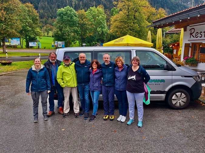 Group of eight people in front of a silver van with mountain scenery in the background.