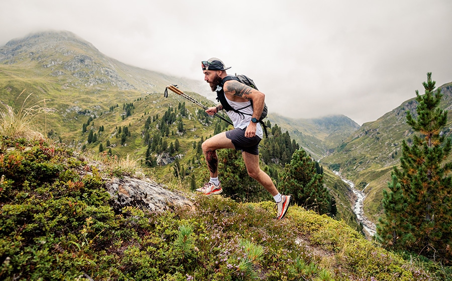 Coureur de trail sur un sentier alpin dans un paysage montagneux avec vallée, ruisseau et pentes verdoyantes.