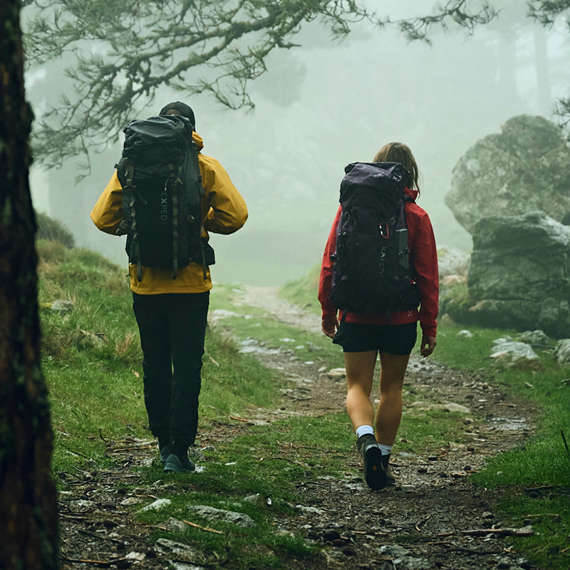 Two people, a man wearing a yellow rain jacket and a large hiking backpack, and a woman wearing a red rain jacket and a hiking backpack, walking through a misty forest landscape.