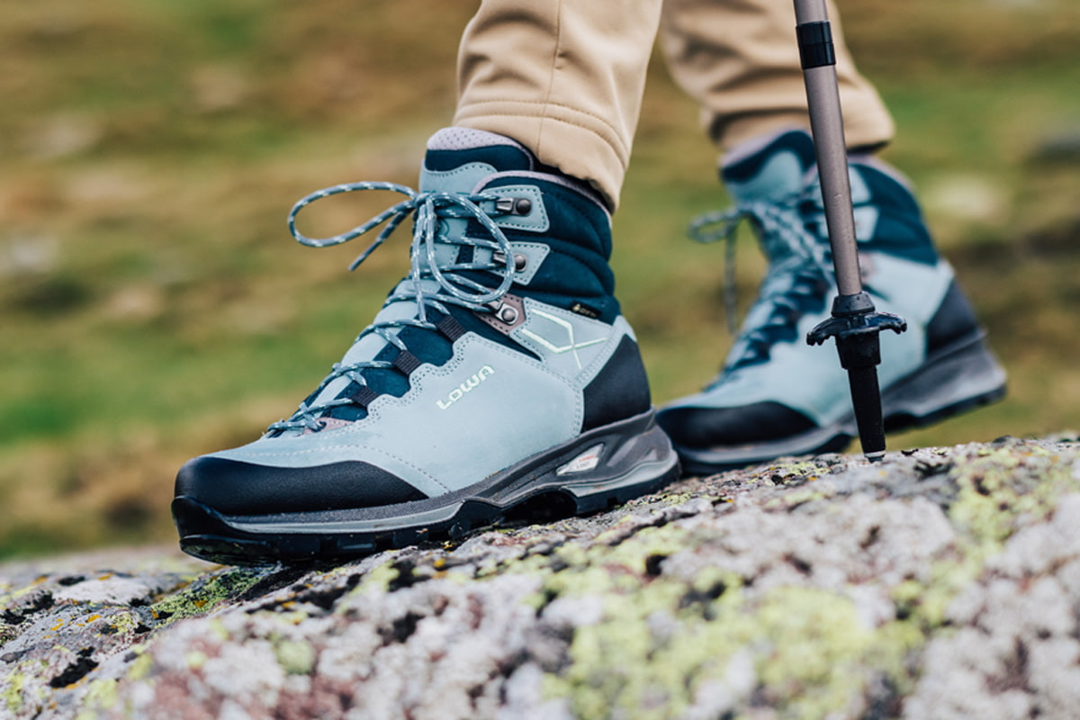 Nahaufnahme eines blauen LADY LIGHT EVO GTX Ws am Fuß einer Wanderin mit Trekkingstöcken auf felsigem Untergrund.