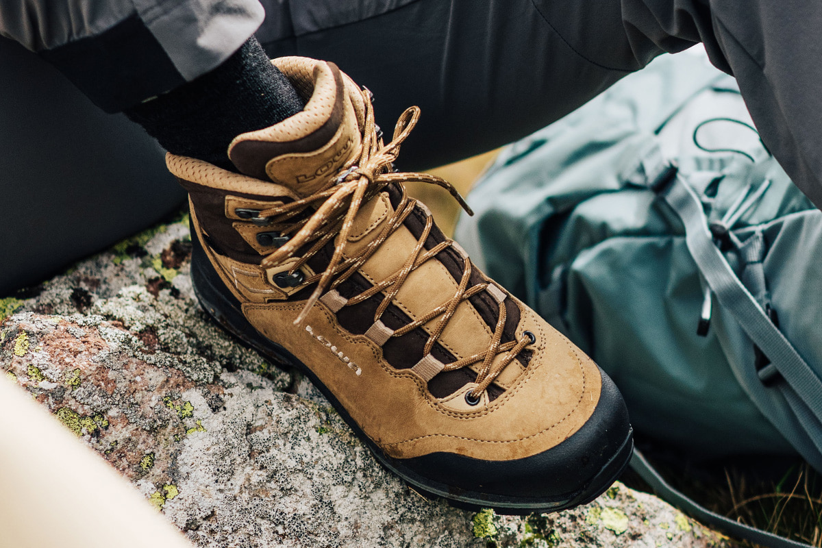Nahaufnahme eines blauen LADY LIGHT EVO GTX Ws am Fuß einer Wanderin mit Trekkingstöcken auf felsigem Untergrund.