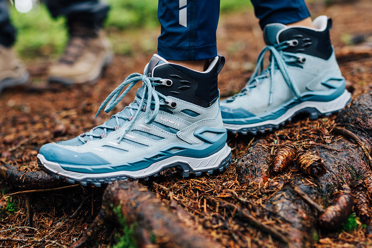 Nahaufnahme eines blauen LADY LIGHT EVO GTX Ws am Fuß einer Wanderin mit Trekkingstöcken auf felsigem Untergrund.